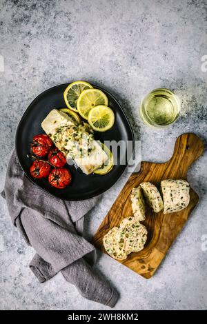 Colpo di testa con filetto di pesce bianco ricoperto di burro composto fuso su un piatto nero con pomodori arrostiti, limoni a fette e vino bianco in un gambo Foto Stock