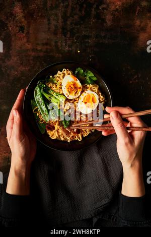 Mani che tengono le bacchette in uovo e tagliatelle vegetali in una ciotola nera con tovagliolo nero Foto Stock