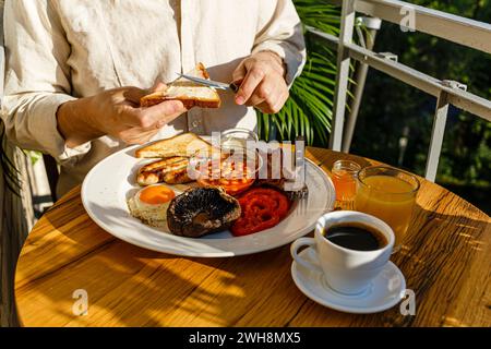 Uomo che mangia la colazione all'inglese completa con uova fritte, salsicce, funghi Portobello, fagioli, toast e caffè serviti su tavola rotonda nella soleggiata estate Foto Stock