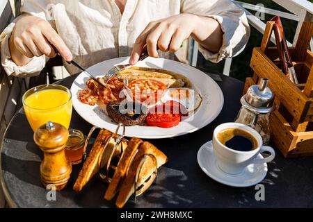 Uomo che mangia la colazione all'inglese completa con uova fritte, salsicce, funghi Portobello, fagioli, toast e caffè serviti su tavola rotonda nella soleggiata estate Foto Stock