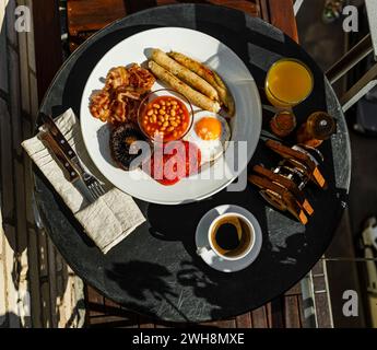 Colazione all'inglese completa con uova fritte, salsicce, pancetta, fagioli, toast e caffè serviti su un tavolo rotondo la mattina d'estate Foto Stock