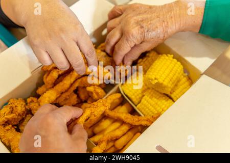 primo piano di tre mani che prendono cibo da una scatola di fast food con pollo, mais, patatine fritte e arepas Foto Stock