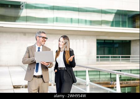 Un uomo d'affari anziano con capelli e occhiali grigi condivide un momento spensierato con una collega più giovane mentre discute di lavoro su un notebook all'aperto, mostrando un tema di mentorship o collaborazione Foto Stock