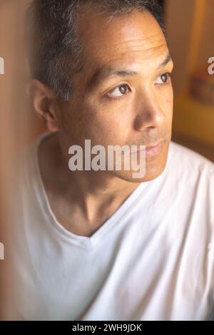 Ritratto verticale di un giovane giapponese che guarda fuori da una finestra con un'espressione pensiva. Concetto relativo alla salute mentale, contemplazione, introspecti Foto Stock