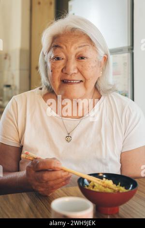 Anziana donna asiatica che mangia riso con bacchette guarda e sorride alla macchina fotografica Foto Stock