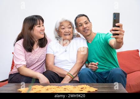 Bambini adulti e anziana madre dai capelli bianchi, famiglia etnica giapponese, scattano un selfie seduto su un divano sorridendo e condividendo un momento di famiglia. Foto Stock