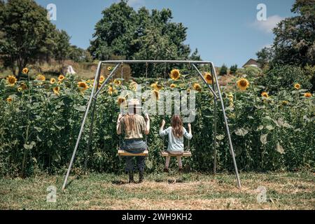 Immagine stravagante di due sorelle su un'altalena che si godono l'estate in un campo di magici girasoli Foto Stock