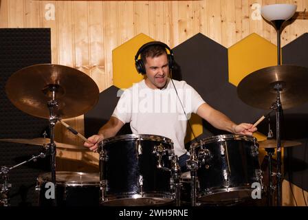 L'uomo bianco caucasico suona emotivamente alla batteria con cuscinetti auricolari in studio sotterraneo. Parete in legno insonorizzata sullo sfondo. Scurire la foto. Musicista Foto Stock