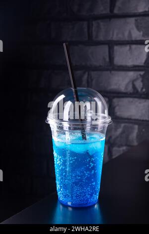 Limonata blu con cubetti di ghiaccio in vetro di plastica decorato con paglia su un bancone di legno contro una parete di mattoni Foto Stock