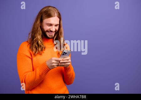 Foto ravvicinata di un giovane e allegro ragazzo di capelli biondi che indossa un maglione arancione che invia messaggi ai suoi amici hipster isolati su uno sfondo di colore viola Foto Stock