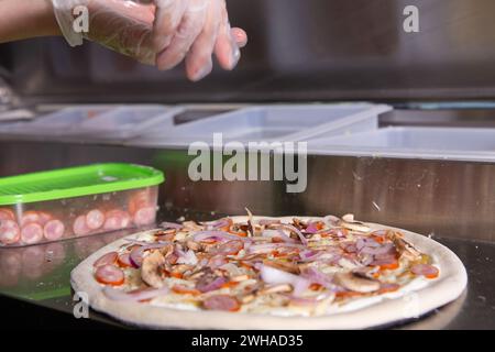 Lo chef prepara una classica pizza italiana con salsicce, funghi e pomodori. Il cuoco cosparge la pizza con i funghi. Foto Stock