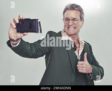 Uomo anziano, moda e selfie con i pollici in alto per un buon lavoro, vincere o fotografare su uno sfondo grigio. Persona matura in stile retrò o vintage Foto Stock