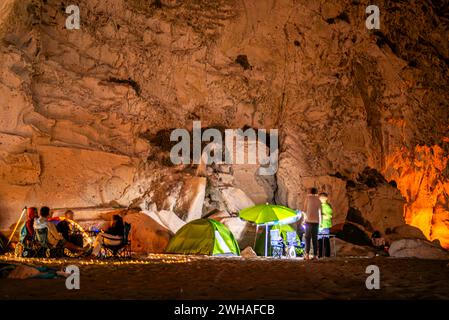 Campeggiatori sotto cieli stellati con tende e luci da campeggio, annidati dal mare e da una montagna rocciosa, creando un ambiente di campeggio costiero sereno Foto Stock