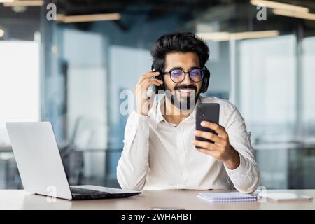 Un uomo con gli occhiali si siede a una scrivania utilizzando un computer portatile e un telefono cellulare, mentre indossa cuffie e occhiali. Ha un sorriso in faccia. Foto Stock