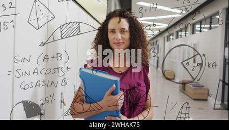 Immagine dei calcoli matematici e della geometria su una felice insegnante birazziale nel corridoio scolastico Foto Stock