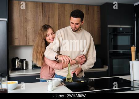 Coppia spagnola sorridente, cucinando omelette e abbracciando in cucina. Foto Stock