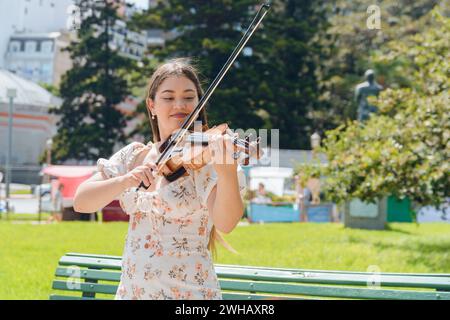 Ritratto in vita di una giovane donna violinista busker latina, con indosso un abito bianco, pelle bianca di etnia venezuelana, felice in piedi giocando all'aperto su pla Foto Stock