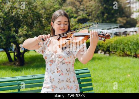 Ritratto in vita di una giovane donna violinista busker latina, con indosso un abito bianco, pelle bianca di etnia venezuelana, felice in piedi giocando all'aperto su pla Foto Stock