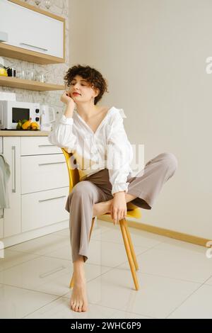 Una giovane donna siede con cura su una sedia gialla in una cucina moderna ben illuminata, che incarna un momento di relax e contemplazione Foto Stock