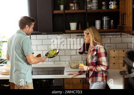 La coppia si impegna in una routine serale rilassata mentre l'uomo cucina con una padella sul fornello mentre la donna, che tiene un drink, guarda in un ben arredato, c Foto Stock