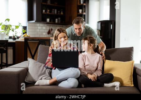 Una famiglia allegra di tre persone trascorre del tempo insieme sul divano del soggiorno, condividendo un momento intorno a un notebook nella loro casa confortevole Foto Stock