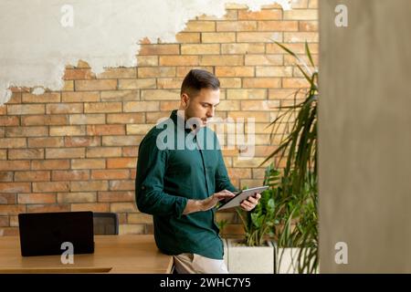 L'uomo allegro sta con sicurezza tenendo un tablet digitale in uno spazio ufficio contemporaneo con una parete in mattoni a vista, a simboleggiare una miscela di tecnologia moderna Foto Stock