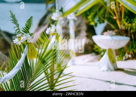 Decorato romantico matrimonio accessori celebrazione sulla spiaggia di sabbia tropicale. Vegetazione lussureggiante e fiori bianchi. Foto Stock