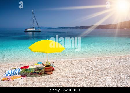 Concetto estivo con accessori in spiaggia. Ombrello giallo su pittoresco sacchetto di paglia di ciottoli, asciugamano, isola di Cefalonia, Grecia. Concetto di vacanza relax in viaggio. Foto Stock