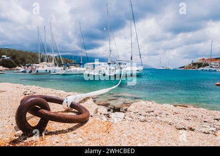 Fune di ormeggio legata ad un anello arrugginito per equipaggiare yacht in background. Splendida vista sul porto di Fiskardo. Pittoresco paesaggio marino del Mar Ionio. Scenario all'aperto dell'isola di Cefalonia, della Grecia, dell'Europa. Foto Stock