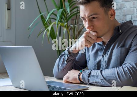 Uomo d'affari premuroso che tocca il mento, riflette idee o strategie, siede alla scrivania di legno con un computer portatile, lavora come libero professionista su progetti online, stud Foto Stock