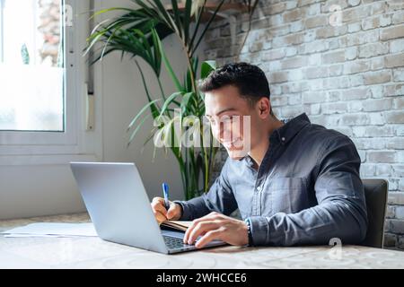 Vista laterale bel giovane uomo d'affari che lavora con il computer in remoto, seduto al tavolo di legno in ufficio. Uomo felice e piacevole che comunica sui social network, cerca informazioni online. Foto Stock