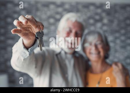 Primo piano di una vecchia coppia caucasica felice mostra le chiavi di un appartamento o di una casa. Un uomo e una donna in pensione sorridenti festeggiano il trasferimento in una nuova casa. Trasloco, affitto, concetto immobiliare. Foto Stock