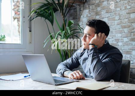 Studente o lavoratore di sesso maschile stanco sedersi a casa scrivania ufficio guardare in lontananza avendo sonno privazione, pigro millenario uomo distratto dal lavoro sentirsi pigro mancanza motivazione, pensando di lavoro monotono opaco Foto Stock