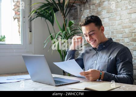 Un uomo d'affari entusiasta studente che legge la lettera postale felicissimo per le grandi notizie, un vincitore maschio felice che tiene in mano il conto cartaceo con approvazione del prestito celebra il rimborso delle tasse ricevere il pagamento dell'aumento dello stipendio seduto alla reception Foto Stock