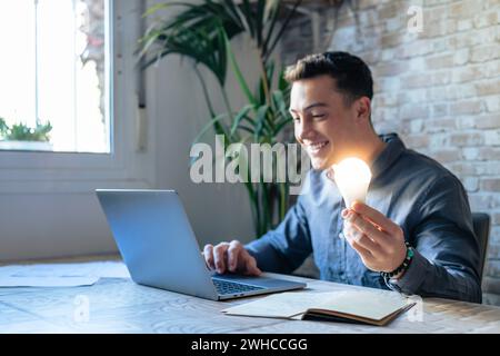 Primo piano del lavoro di un dipendente maschio al computer con una lampadina luminosa brillante ottenere un'idea di business brillante. L'uomo d'affari o il lavoratore si sentono motivati dal successo del progetto di avvio in ufficio. Concetto di innovazione. Foto Stock