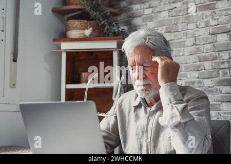 Stanco uomo maturo degli anni '70 che lavora al notebook da casa, togliendo gli occhiali, toccando la testa e gli occhi. Vecchio pensionato maschio che soffre di mal di testa, vertigini dopo aver guardato lo schermo del computer. Problema alla vista Foto Stock