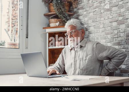 Uomo anziano di mezza età dai capelli grigi seduto da solo in cucina su una sedia che si allunga dopo molto tempo lavorando al computer facendo esercizi per i lombini che prevengono l'osteoartrite e la stenosi spinale Foto Stock