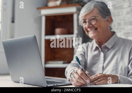 Lavoratore remoto anziano. Una donna anziana concentrata negli occhiali lavora su un computer portatile dall'ufficio domestico leggendo i documenti elettronici delle e-mail. Lavoratrice freelance anziana siede al tavolo della cucina per pc scrivendo online Foto Stock