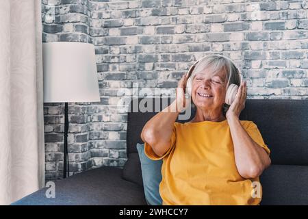 Vista laterale felice vecchia donna in pensione matura che ascolta musica popolare con le cuffie, rilassati da solo su un comodo divano, goditi un weekend tranquillo e spensierato Foto Stock