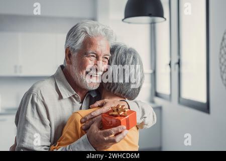 Una donna anziana sorridente che regala una confezione regalo al vecchio marito sorridente degli anni '80, congratulandosi con il compleanno o l'anniversario del matrimonio. Un uomo anziano gioioso che si sente entusiasta di ricevere un regalo dalla moglie Foto Stock