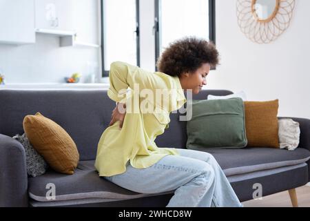 La studentessa afro soffre di mal di schiena che si sente male, seduta sul divano nel salotto di casa. Ha un dolore alla schiena dopo aver studiato al computer per molto tempo. Concetto di lavoro sedentario e cattiva postura Foto Stock