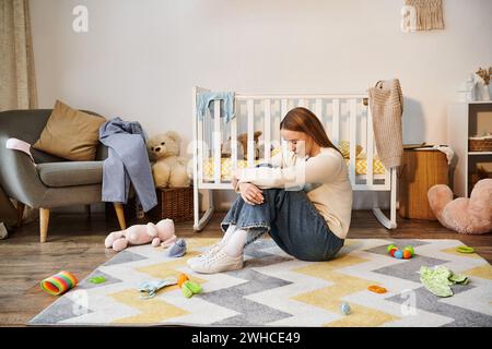 giovane donna sconfitta seduta sul pavimento con la testa inchinata vicino ai giocattoli e alla culla nella stanza del letto di casa Foto Stock