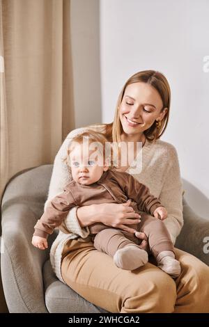 donna allegra e attraente seduta in poltrona con bambino in un'accogliente stanza da letto, madre e figlio Foto Stock