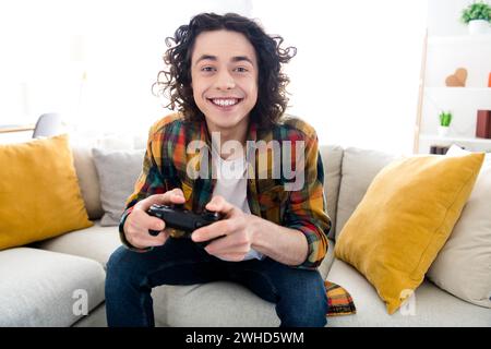 Foto di un ragazzo allegro e divertente che indossa una camicia a quadri mentre gioca a playstation all'interno della casa Foto Stock