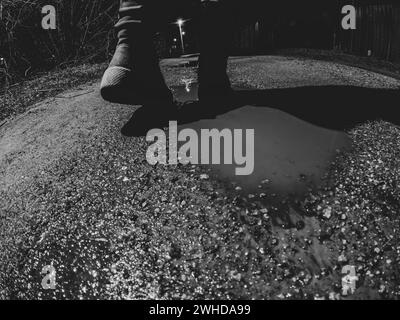 Una ragazza cammina lungo una bagnata strada invernale fino a casa al buio, vista dal basso verso l'alto, foto in bianco e nero, piedi sulla strada al buio Foto Stock