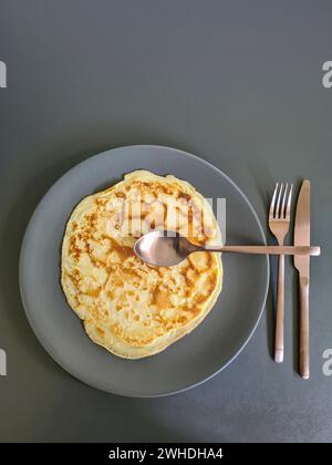 Pancake su piatto grigio con coltello da posate, forchetta e cucchiaio davanti allo sfondo grigio Foto Stock