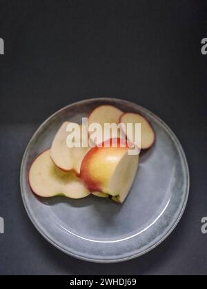 Una mela rossa e verde giace a fette su un piatto di colore chiaro con sfondo scuro Foto Stock