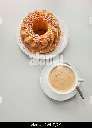 Una tazza di caffè bianco con piattino e cucchiaio è elegantemente disposta accanto a una torta colorata su un tavolo bianco Foto Stock