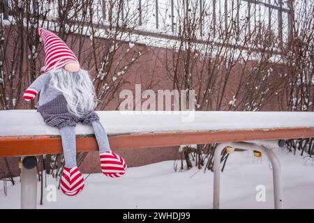 Nano sulla panchina nella neve. Gnomo solitario. GNOME nella neve. Sfondo d'amore. Nano da favola natalizia. Decorazioni per le vacanze invernali. Adorabile bambola nana. Foto Stock