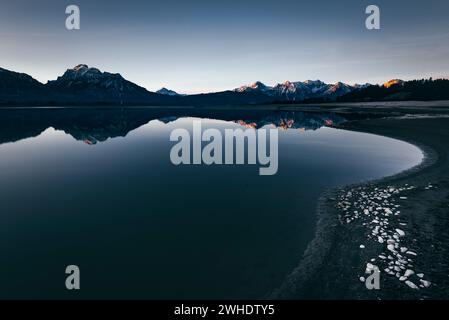 Panorama alpino con riflessi nel Forggensee, parzialmente drenato, vicino a Füssen nel Allgäu. Alpenglow alla prima luce del sole. In primo piano, pietre sul letto del lago ghiacciato. Foto Stock